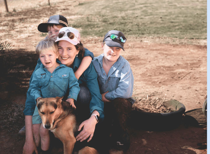 A woman, her children hug and smile, with their dog, to a backdrop of rural Australia