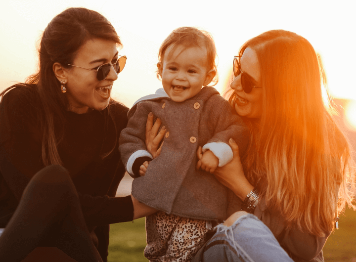 A toddler smiles as two women smile encouragingly at her in their arms