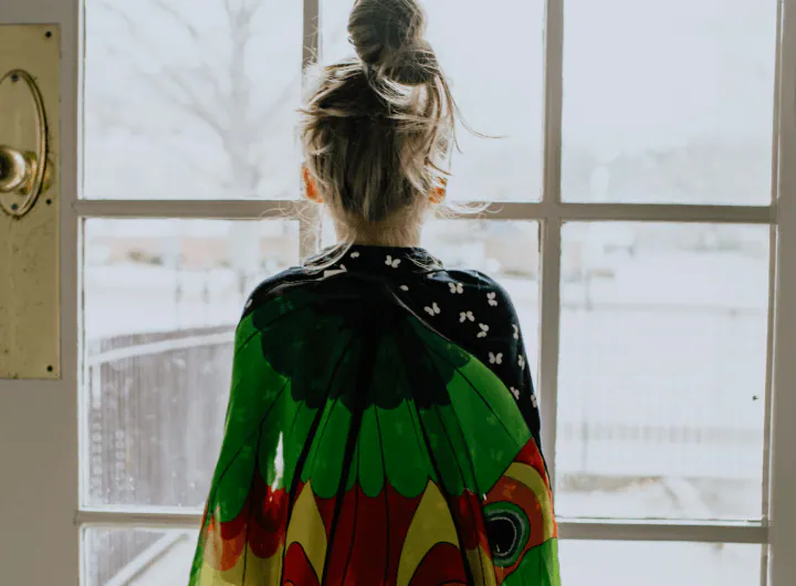 A young girl wears a butterfly cape. She has her back to us and is looking out a door.