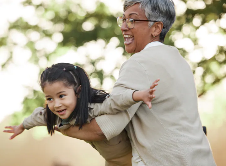 A grandmother joyfully playing with her granddaughter, lifting her in the air like an airplane in an outdoor setting.