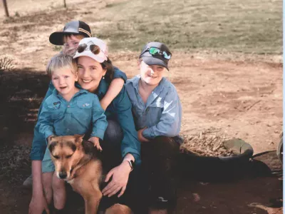 A woman, her children hug and smile, with their dog, to a backdrop of rural Australia