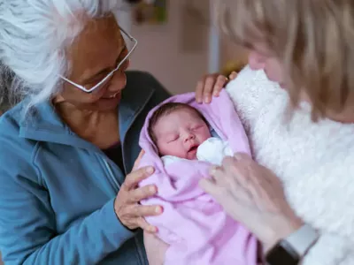 A swaddled baby sleeps, as two older women look on, providing support to the new parent.